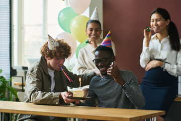 Group of happy young intercultural colleagues in birthday caps whistling in noisemakers while one...