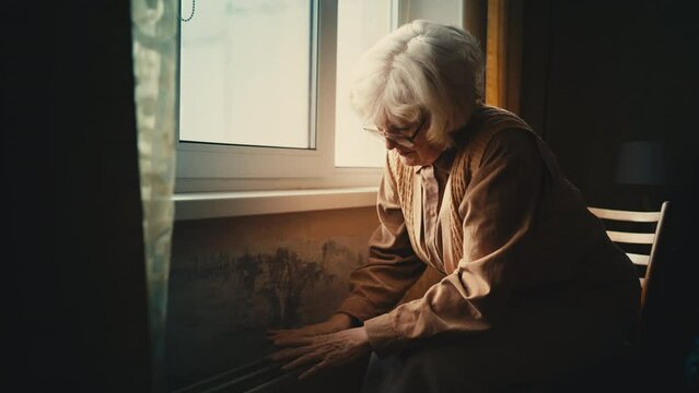 Poor elderly woman warming her hands near radiator, low temperature in a room