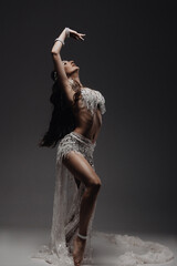 Belly and body of a woman dancing oriental dance in the studio, making beautiful body movements, she is dressed in a chic white suit on a dark background of the studio - obrazy, fototapety, plakaty