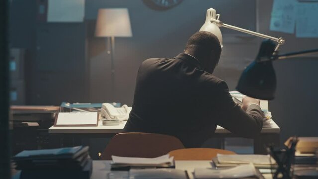 Back view of African American businessman in suit sitting at desk in dark office, reading paper documents and catching deadline alone at night