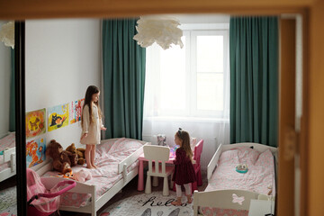 Adorable girl in white dress or night gown standing on bed with toys and looking at her cute sister...