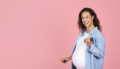 Smiling pregnant brunette lady exercising with barbells on pink background
