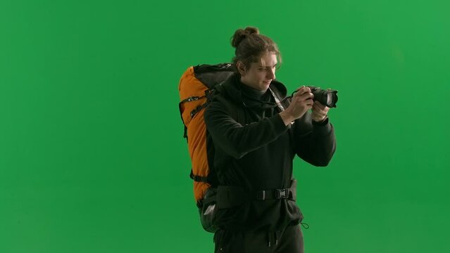A male traveler takes pictures with a camera while hiking. A tourist with a backpack on his back stands in a studio on a green screen close up. Concept of travel, active rest, hiking.
