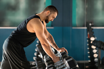 Sport Equipment Concept. Young Male Athlete Taking Dumbbell From Rack At Gym