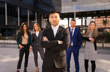 serious asian executive arms crossed with employees behind multiethnic group - obrazy, fototapety, plakaty