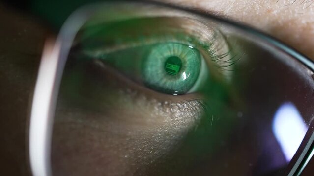 Blurred vision of a computer screen with code reflecting on the surface of eyeglasses. Close-up of a human eye reflecting green coding text, seen through the lens of glasses