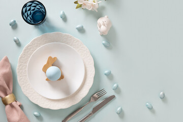 Easter table setting with white plate, eggs and bunny on blue background. View from above. Space for greeting text.
