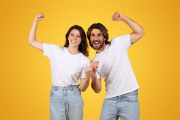 Enthusiastic young couple in white t-shirts and jeans cheerfully raising fists in a victorious gesture