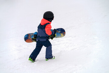 A small child with a snowboard under his arm goes uphill at a ski resort. Copy space.
