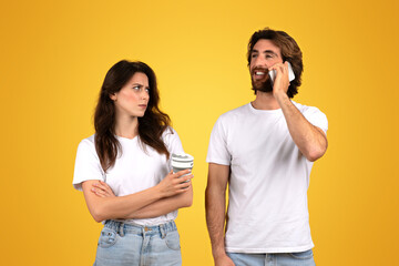 Skeptical woman holding a coffee cup looking at a smiling man talking on a mobile phone