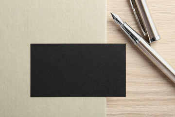 Blank black business card, fountain pen and notebook on wooden table, top view. Mockup for design