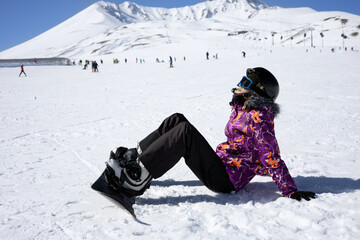 Girl or boy in ski helmet, sunscreen mask and balaclava sits on snow with snowboard and relaxes against the backdrop of snow-covered mountain ski slope and a cloudy sky. Winter. Sport and travel 