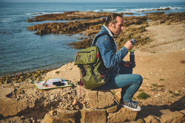 Full length portrait of a handsome hiker traveler man sitting on a rock by sea, drinking hot drink. People Travel Nature