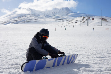 Girl or boy in ski helmet, sunscreen mask and balaclava sits on snow and fix the snowboard bindings against the backdrop of snow-covered mountain ski slope and a cloudy sky. Winter. Sport and travel  - obrazy, fototapety, plakaty