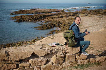 Full length portrait of a handsome traveler man in casual denim, drinking hot drink while taking a coffee break by sea