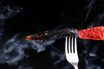 Burnt dried red hot pepper on a fork in smoke on a black background