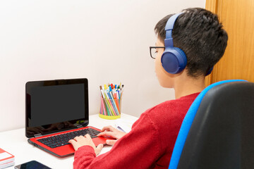 Young student using a laptop for homework, wearing wireless headphones to attend online classes in...