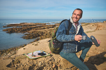 Happy male tourist sitting on a rock by sea, holding a thermos bottle and pouring some coffee into a steel mug