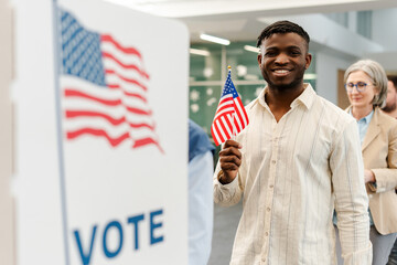 Portrait of smiling African American man holding American flag vote at polling station. Democracy,...