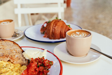 Scrambled eggs, toasted bread, tomatoes, pancakes with berries, cappuccino - breakfast on a white table in a cafe 