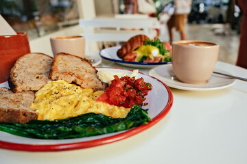 Scrambled eggs, toasted bread, tomatoes, croissant with eggs, cappuccino - breakfast on a white table in a cafe 