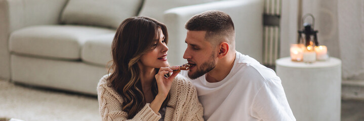 Banner. Beautiful young loving couple having breakfast, drinking hot chocolate or coffee with marshmallow in bed at home. Happy spouses enjoying lazy romantic winter morning in bedroom. Candles