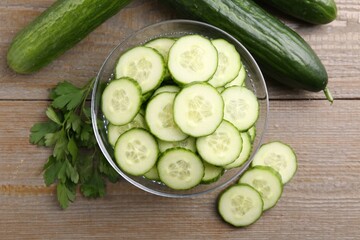 Cut cucumber in glass bowl, fresh vegetables and parsley on wooden table, flat lay
