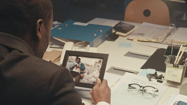 Over shoulder of busy African American businessman looking at photo of his kids in frame and missing them while overworking in office late at night