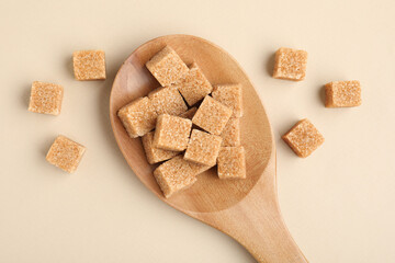 Brown sugar cubes in wooden spoon on beige background, top view
