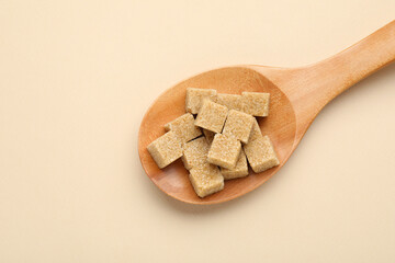 Brown sugar cubes in wooden spoon on beige background, top view