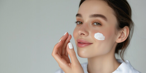 Smiling young woman applying facial cream. Close-up beauty portrait with skincare product on cheek, light grey background. Skincare and wellness concept. Design for banner with copy space or cosmetic.