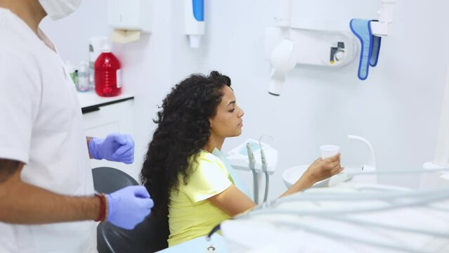 Young latin woman patient rinse her mouth over a dental cuspidor while her dentist is waiting to continue procedure. Young female patient rinses her mouth after treatment at a modern dental clinic