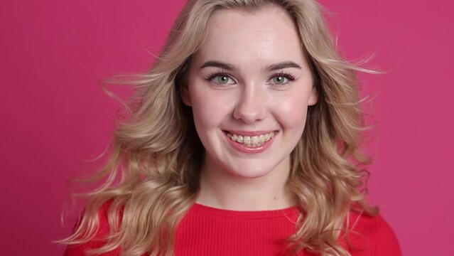 Beautiful young caucasian woman with transparent ceramic braces on teeth isolated on pink backdrop. Girl smiles, looks at camera, blond curly hair moves from fan. Beauty, health and fashion concept.