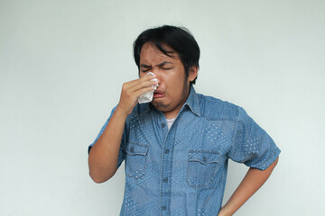 A young Asian man in a blue shirt sneezes and holds a tissue. Flu illness concept. Isolated in gray...
