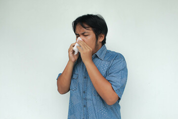 A young Asian man in a blue shirt sneezes and holds a tissue. Flu illness concept. Isolated in gray...