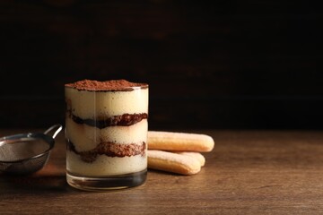 Delicious tiramisu in glass, biscuits and sieve on wooden table. Space for text