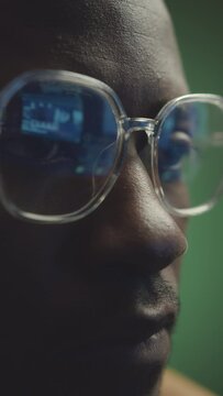 Vertical closeup of focused young Black male programmer with computer data reflecting in his eyeglasses working at night