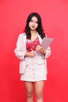 Portrait Of A Young Asian Indonesian Woman In An Office Wearing A Cream-colored Blazer Carrying A Tablet + Pen. Isolated Red Background For Content Materials, Banners, Advertising.