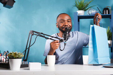 Vlogger announcing giveaway and showing shopping bag with gifts while broadcasting live. Smiling man blogger speaking to subscribers, promoting product and looking at camera