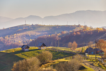 Panoramic view of misty sunny autumn evening in mountains. Beautiful sunset hills landscape. Slopes, meadows, fields, village, house, dirt road. Amazing fall rural scene. Carpathian range. - 724265840