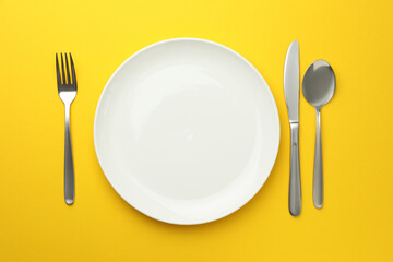 Clean plate, fork, knife and spoon on yellow background, flat lay