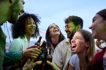 Cheerful multiracial young people toast with beer bottles gathered celebrating laughing on summer outdoors. Excited group happy millennial friends together enjoying sunset drinks at rooftop party  - obrazy, fototapety, plakaty