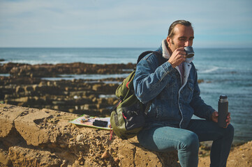 Handsome hiker traveler man sitting on the cliff and drinking hot drink