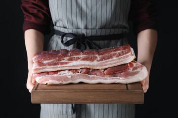 Woman holding wooden board with pieces of raw pork belly on black background, closeup