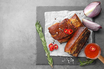 Pieces of baked pork belly served with sauce, rosemary and chili pepper on grey table, top view. Space for text