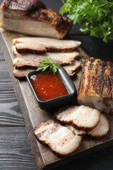 Pieces of baked pork belly served with sauce and parsley on black wooden table, closeup