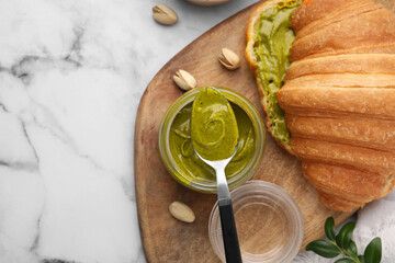 Tasty pistachio cream in jar, spoon and croissant on white marble table, top view. Space for text