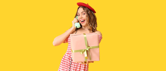 Young woman with gift talking by retro telephone on yellow background. International Women's Day