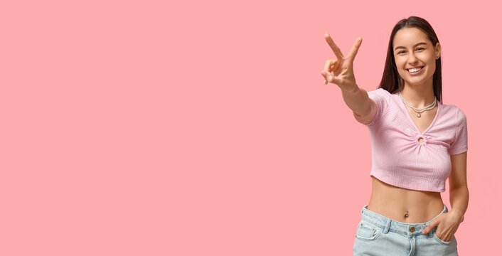 Sexy young woman with navel piercing showing victory gesture on pink background with space for text