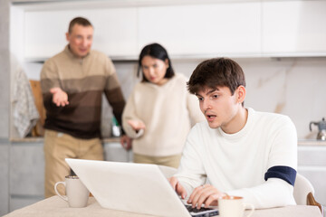 Young woman and adult man quarreling with young guy working on laptop in kitchen at home..